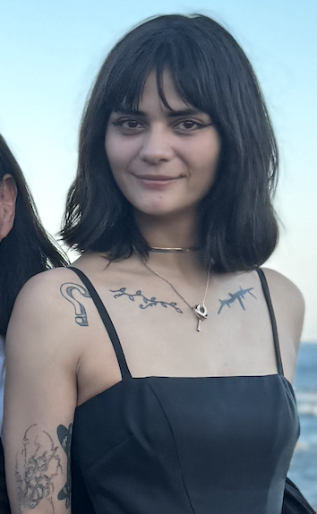 Violet Benjamin — outdoors by the water, shoulder-length black hair with bangs, black top, gold choker necklace, visible tattoos: thorny branch design across chest and collarbone, ear tattoo on right upper arm, line art tattoos on left shoulder, abstract designs on right forearm